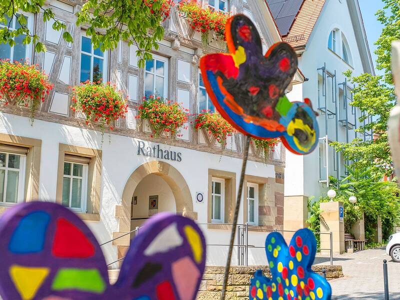 Vor dem Fachwerk-Rathaus, geschmückt mit roten Blumen, stehen farbenfrohe, abstrakte Kunstwerke auf Stäben.