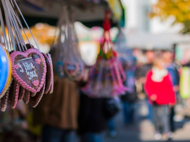 Im Vordergrund hängt ein Lebkuchenherz mit der Aufschrift "I like you" neben bunten Ringen, und im Hintergrund bewegt sich eine unscharfe Menschenmenge.
