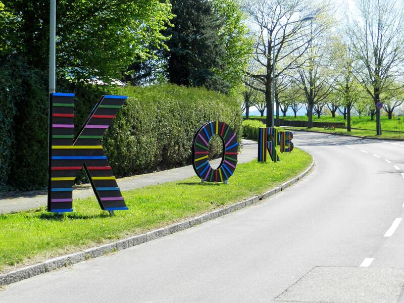 Am Straßenrand stehen große, bunte Buchstaben "K", "O", "B" und "E" aus horizontalen Streifen in verschiedenen Farben, umgeben von grüner Vegetation.