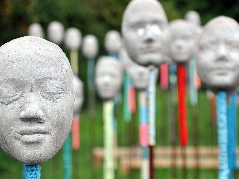 Eine Gruppe von grauen Skulpturen eines menschlichen Gesichts auf bunten Stangen steht in einem Grünen Hintergrund.