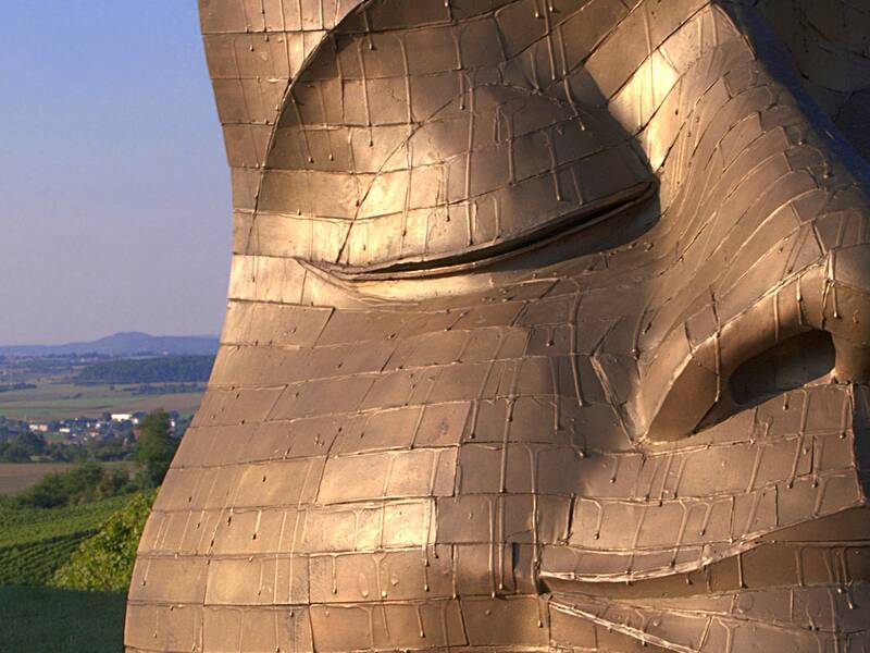 Nahaufnahme einer großen, metallischen Skulptur eines Gesichts mit geschlossenen Augen, im Hintergrund eine weite Landschaft mit Feldern und Hügeln.