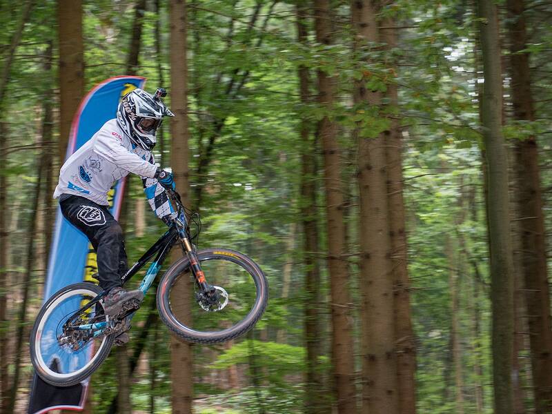 Eine Person mit Helm und Schutzkleidung springt mit einem Mountainbike in einem Wald über eine Rampe.