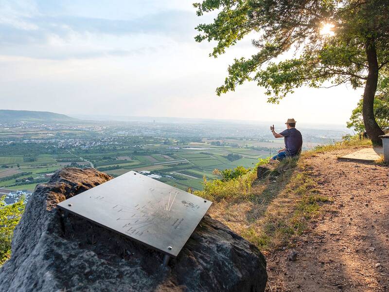 Ein Mann sitzt auf einem Hügel mit Blick auf die weite Landschaft und macht ein Foto, während die Sonne durch die Bäume scheint.