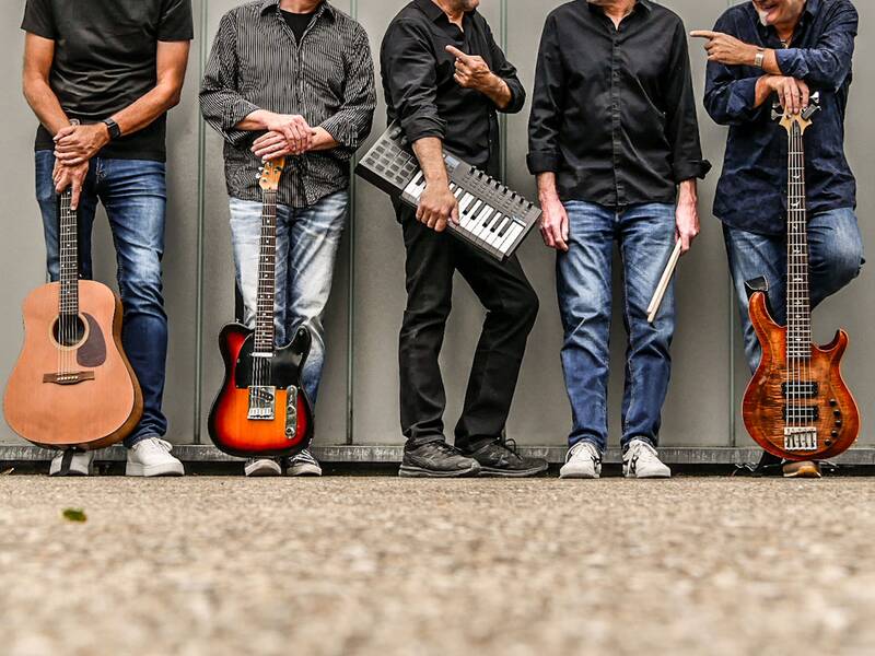 Fünf Männer stehen nebeneinander vor einer grauen Wand und halten Musikinstrumente, darunter Gitarren, ein Keyboard und ein Drumstick.