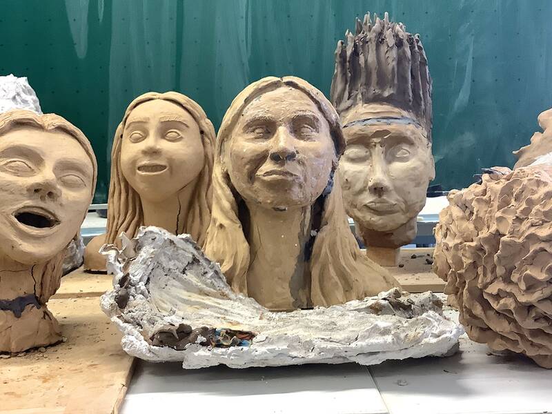 Tonbüsten mit verschiedenen Gesichtsausdrücken und Frisuren stehen in einer Werkstatt auf einem Tisch, umgeben von Gipsformen.