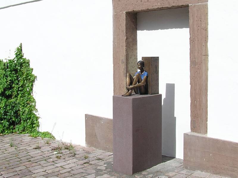 Eine Bronzeskulptur eines sitzenden Mädchens mit blauem Oberteil steht auf einem Sockel in einem Rahmen an einer weißen Wand neben einer Begrünung.