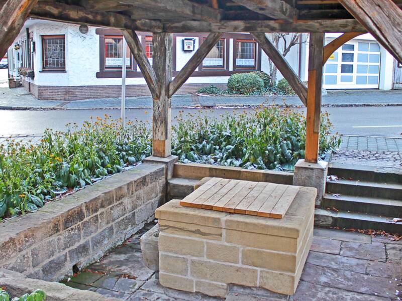 Ein überdachter Brunnen mit Holzbalken steht umgeben von niedrigen Steinmauern und Treppen, während im Hintergrund eine Straße und ein Fachwerkgebäude zu sehen sind.