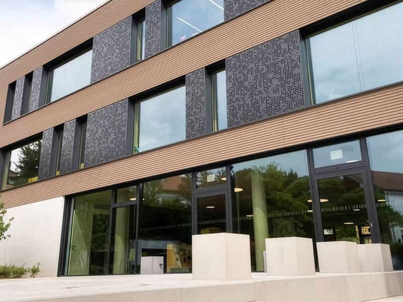 Das Bild zeigt ein modernes Gebäude mit großen Glasfenstern und einer Fassade aus horizontalen Holzpaneelen sowie dekorativen schwarzen Elementen.