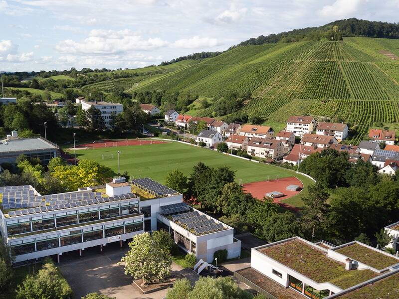 Eine Schule mit Solaranlagen auf dem Dach liegt inmitten einer grünen Landschaft mit Weinbergen und neben einer Sportanlage, umgeben von Wohnhäusern und üppiger Vegetation.