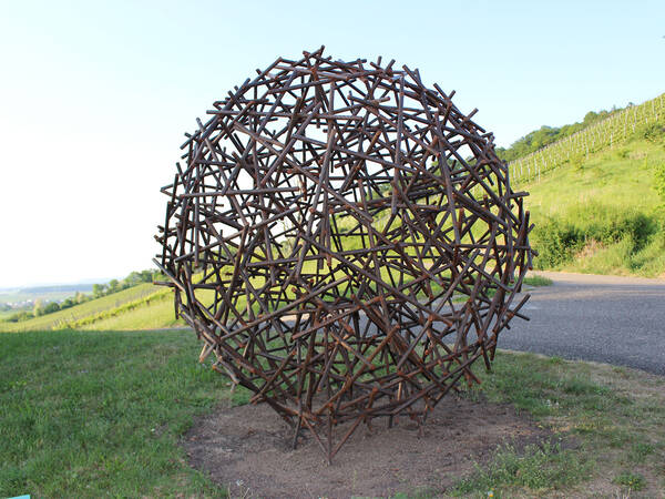 Eine kugelförmige Skulptur aus ineinander verflochtenen Holzästen steht auf einer grasbewachsenen Fläche neben einer Straße, umgeben von grünen Hügeln.