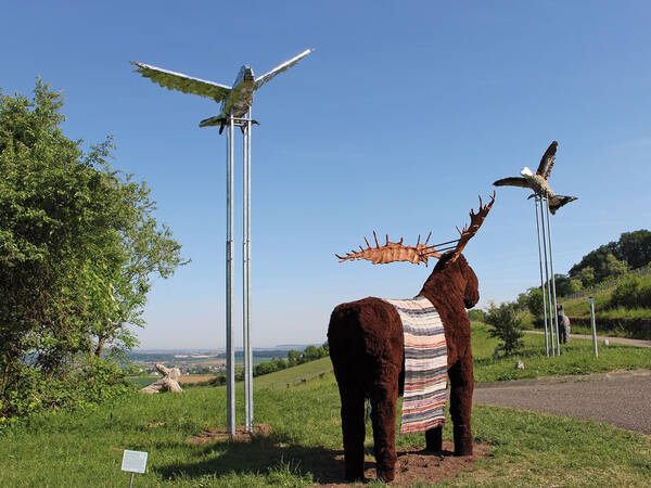 Eine Skulptur eines Elches mit einem gestreiften Schal steht auf einer Wiese, während im Hintergrund Metallvögel in den Himmel ragen.