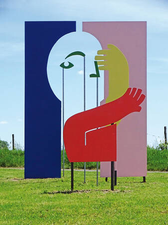 Eine bunte, abstrakte Skulptur mit grafischen Elementen in Form von Armen und einem Gesicht steht auf einer Wiese vor einem blauen und einem rosa Hintergrund.