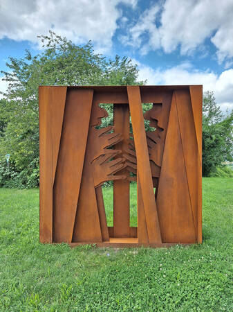 Eine rostfarbene Metallskulptur mit ausgeschnittenen Silhouetten von zwei Profilen und Händen steht auf einer Wiese vor einem Hintergrund aus Bäumen und wolkenbedecktem Himmel.