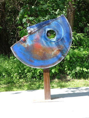 Ein farbenfrohes, abstraktes Kunstwerk in Form einer Scheibe mit einem Loch in der Mitte steht auf einem Holzpfosten, umgeben von Grünpflanzen.