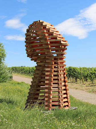 Eine skulpturale Struktur aus gestapelten Holzblöcken erhebt sich auf einer Wiese neben einem Weg, umgeben von einem Weinberg unter einem blauen Himmel.