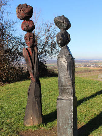 Zwei abstrakte Holzskulpturen, die menschliche Figuren mit Gesichtskonturen darstellen, stehen auf einem grünen Feld vor einem klaren blauen Himmel.