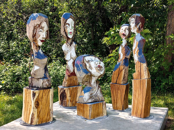 Fünf aus Holz geschnitzte Figuren mit bemalten Details stehen auf Sockeln im Freien, umgeben von grüner Vegetation.