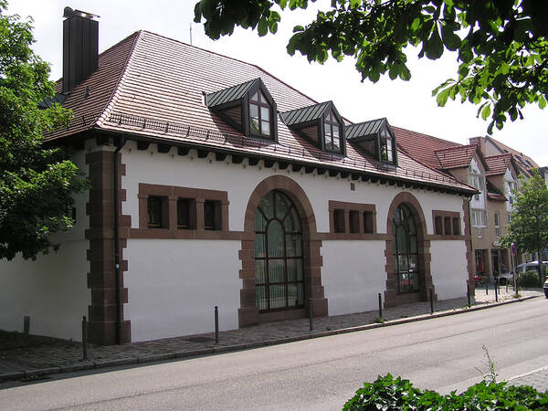 Ein Gebäude mit roten Dachziegeln und großen, gewölbten Fenstern steht an einer ruhigen Straße, umgeben von Bäumen.
