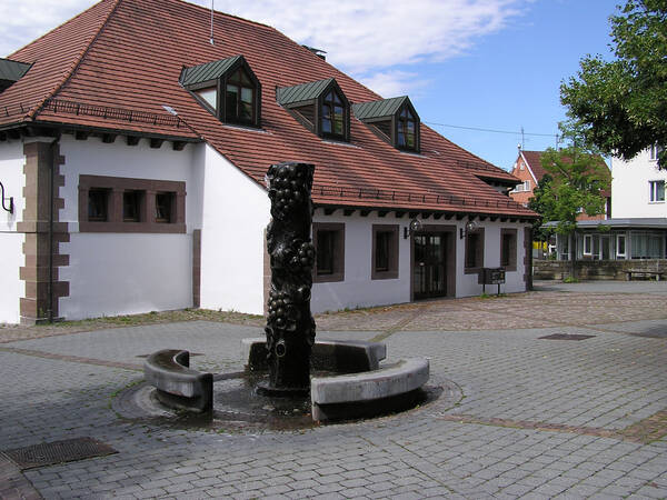 Ein kleiner Platz mit einem skulpturalen Brunnen in der Mitte, umgeben von einem Gebäude mit einem roten Ziegeldach und mehreren Dachgauben.