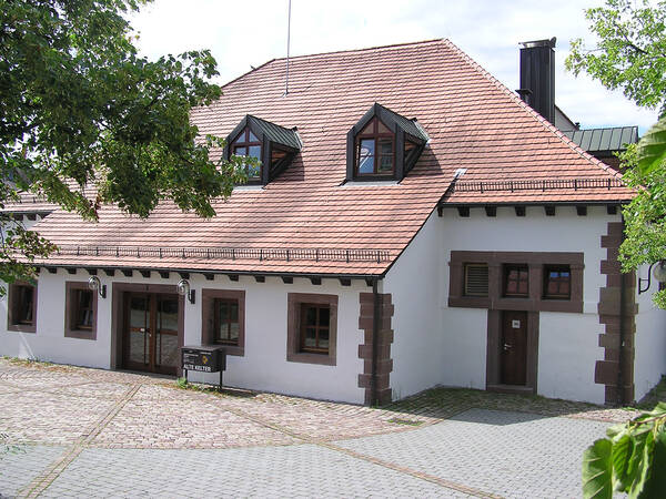 Ein traditionelles Gebäude mit weißer Fassade und rotem Ziegeldach steht auf einem gepflasterten Platz, umgeben von Bäumen.