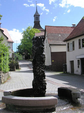 Ein kunstvoll gestalteter Brunnen steht im Vordergrund eines gepflasterten Platzes, umgeben von traditionellen Gebäuden mit einem Kirchturm im Hintergrund.