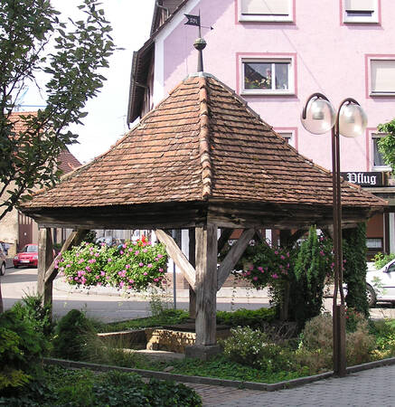 Ein überdachter, aus Holz gebauter Brunnen steht in einem kleinen begrünten Platz, umgeben von Blumen und einem Gebäude im Hintergrund.