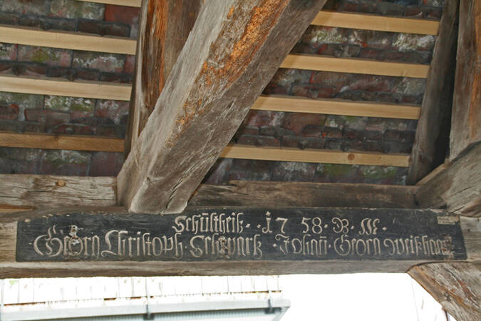 Ein Holzbalken in einem Fachwerkgebäude zeigt eine Inschrift in alter Schriftart, umgeben von sichtbaren Dachziegeln und Holzelementen.