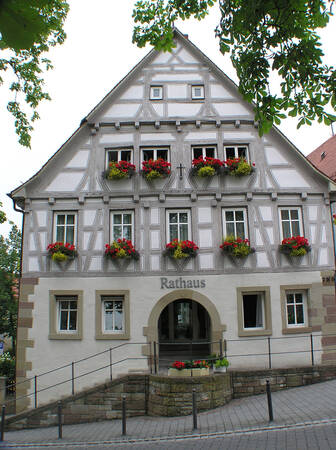 Das Bild zeigt ein traditionelles Fachwerk-Rathaus mit mehreren Fenstern, die mit roten und gelben Blumen geschmückt sind, und einem Eingang mit Treppe.