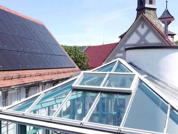 Ein Gebäude mit einem Glasdach im Vordergrund, einer großen Solaranlage auf dem Dach links und einem kirchenähnlichen Turm im Hintergrund.