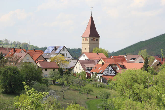 Eine ländliche Landschaft mit einem historischen Kirchturm, der von traditionellen Häusern mit roten Dächern umgeben ist, eingebettet in viel Grün.