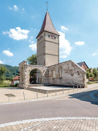 Ein historischer Kirchturm mit rotem Dach und zwei Zifferblättern steht neben den Ruinen eines alten Gebäudes unter einem blauen Himmel mit einigen Wolken.