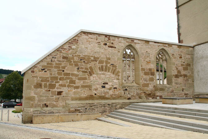 Eine alte Steinmauer mit zwei gotischen Fenstern und kleinen Treppen im Vordergrund.