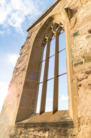 Ein gotisches Fenster in einer alten Steinmauer ragt vor einem blauen Himmel mit einigen Wolken empor.