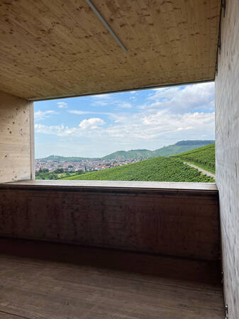 Ein rechteckiges Fenster aus Holz bietet den Blick auf eine hügelige Landschaft mit Weinbergen und einem blauen Himmel.
