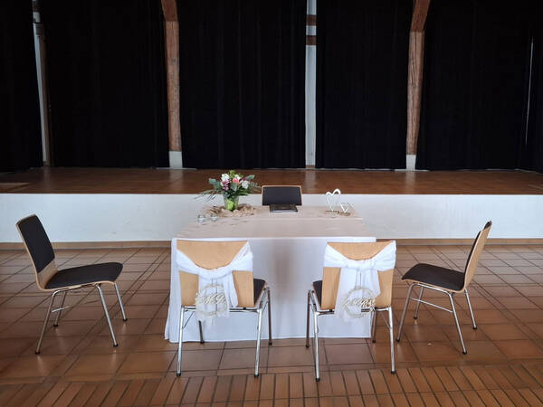 Ein mit weißer Tischdecke gedeckter Tisch steht vor einer Bühne mit schwarzem Vorhang, umgeben von vier Stühlen, und ist mit einem Blumenarrangement geschmückt.