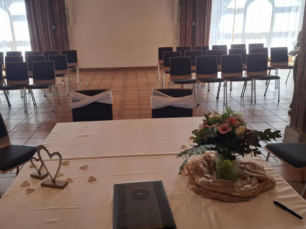 Ein Veranstaltungsraum mit Reihen von Stühlen, einem dekorierten Tisch mit Blumen, einem Buch und einem Herzornament.