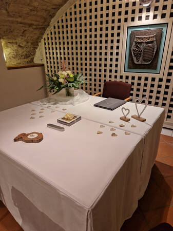 Ein eleganter, mit weißem Tuch gedeckter Tisch ist dekoriert mit Blumen, herzförmigen Ornamenten und einer Tafel mit der Aufschrift "KORB" an der mit Stein dekorierten Wand dahinter.