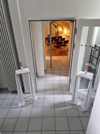Ein heller Eingangsbereich mit zwei Kerzenständern führt zu einem gemütlichen Raum mit mehreren Stühlen und einem Stativ.