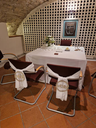Ein elegant dekorierter Tisch mit Blumen und herzförmigem Dekor in einem Raum mit gemustertem Gitterhintergrund und zwei Stühlen, die mit weißen Tüchern verziert sind.