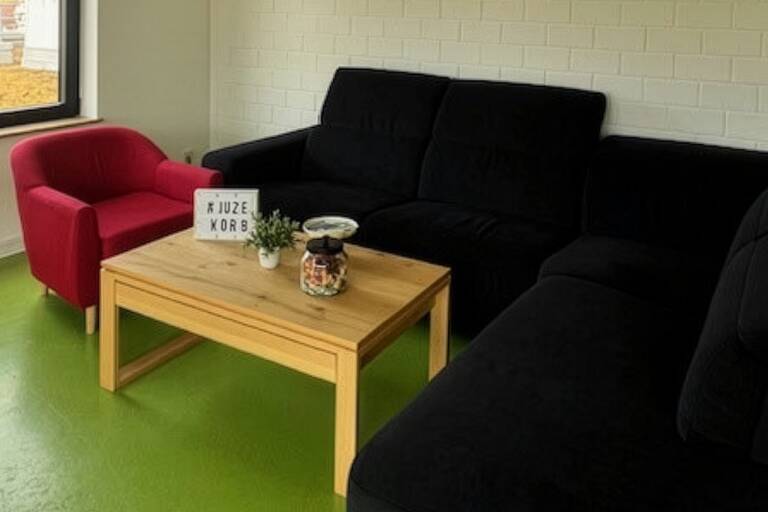 Ein Wohnzimmer mit einem schwarzen Ecksofa, einem roten Sessel und einem hölzernen Couchtisch auf einem grünen Boden.