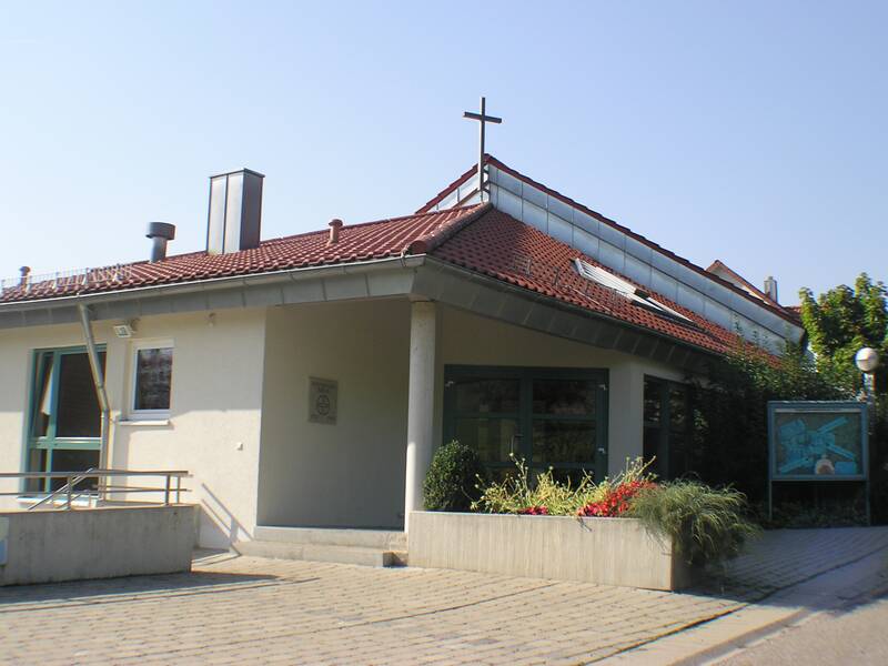 Ein modernes Gebäude mit einem roten Ziegeldach, auf dem ein Kreuz angebracht ist, umgeben von Pflanzen und einem Pflasterweg.