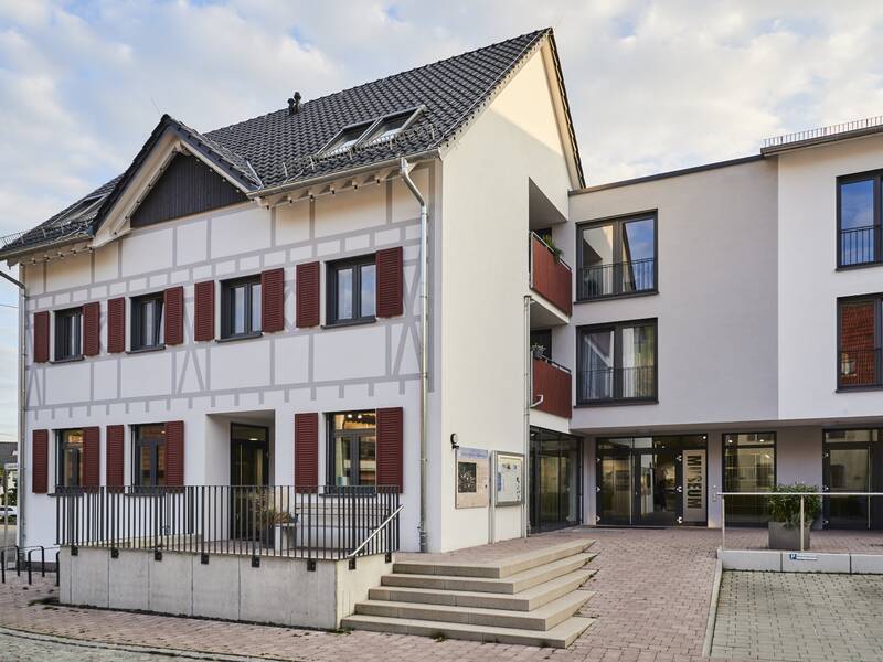 Das Bild zeigt ein modernes Gebäude mit einer Mischung aus traditionellem Fachwerkstil und zeitgenössischer Architektur, inklusive großer Fenster und einem Eingangsbereich mit Treppen.