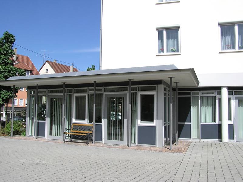 Ein moderner Eingangsbereich mit großen Glasfenstern und einer Bank davor, neben einem großen, hellen Gebäude.