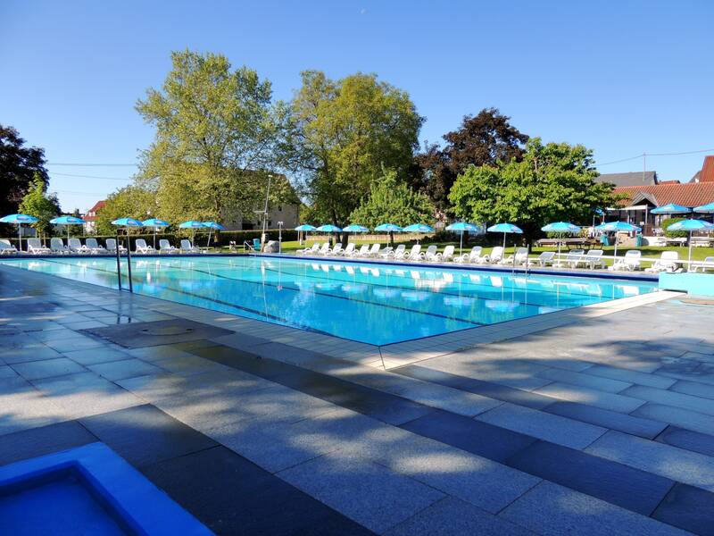 Ein leeres Schwimmbecken ist von Liegestühlen und Sonnenschirmen umgeben, mit Bäumen und Gebäuden im Hintergrund bei klarem Himmel.