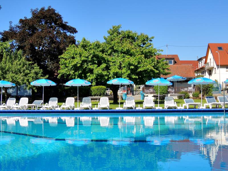Ein Freibad mit klarem Wasser, umgeben von weißen Sonnenliegen und blauen Sonnenschirmen vor einer Reihe grüner Bäume und Gebäude im Hintergrund.