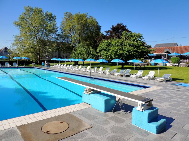 Ein großes Freibad mit einem Sprungbrett, umgeben von Liegestühlen und blauen Sonnenschirmen, steht neben einer Rasenfläche mit Bäumen.