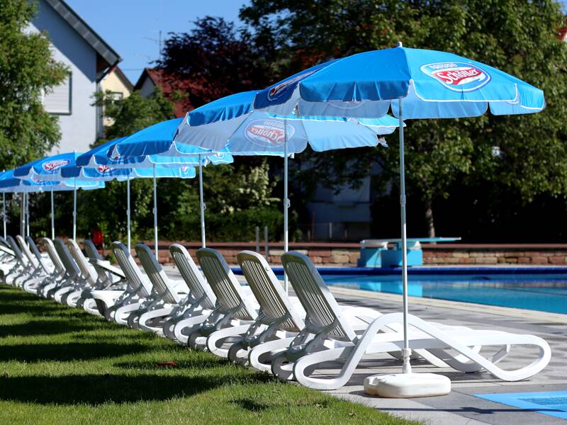 Eine Reihe von weißen Liegestühlen mit blauen Sonnenschirmen steht am Rand eines Schwimmbeckens auf einer Grünfläche.
