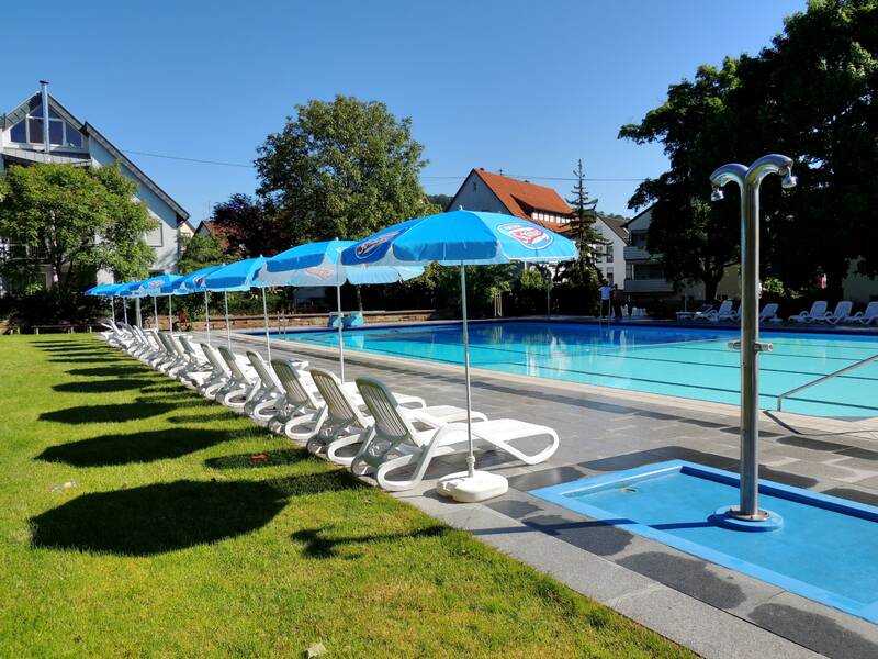 Ein Freibad mit einem großen Pool, umgeben von weißen Liegestühlen und blauen Sonnenschirmen auf einer grasbewachsenen Fläche, bei sonnigem Wetter.