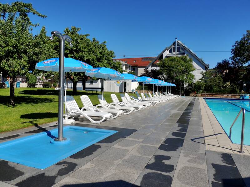 Eine Reihe von weißen Sonnenliegen und blauen Sonnenschirmen steht neben einem Pool auf einer gepflasterten Fläche mit grünen Bäumen im Hintergrund.