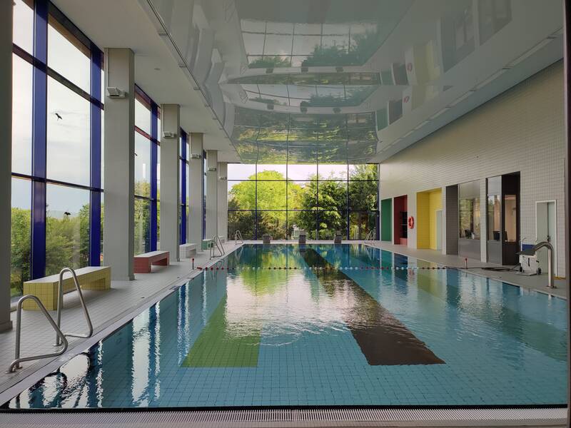 Ein lichtdurchflutetes Hallenbad mit großen Fenstern, durch die man grüne Bäume draußen sieht, wird gezeigt.
